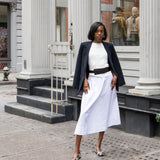 Woman in a white dress and black jacket standing on a city street with classical architecture.