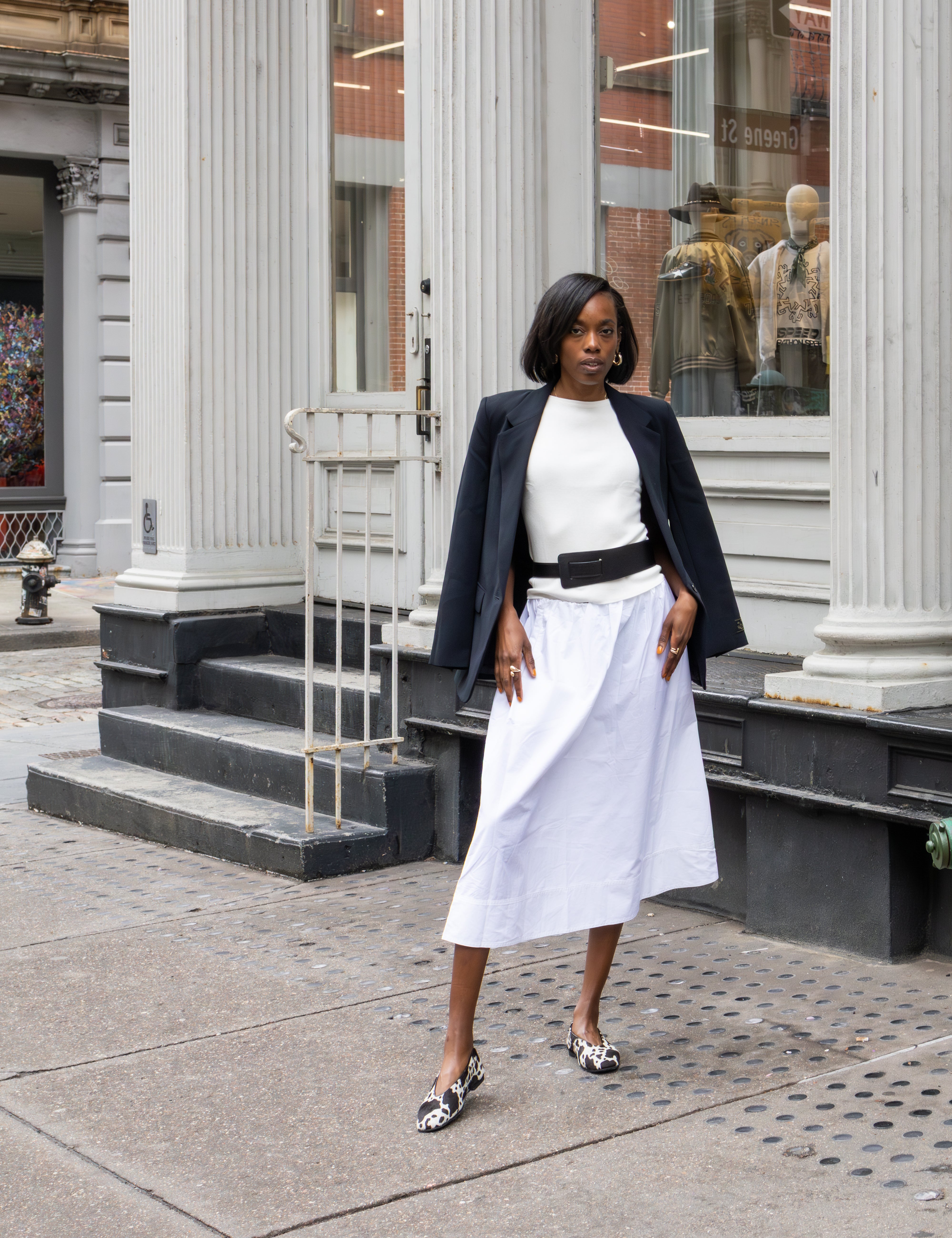 Woman in a white dress and black jacket standing on a city street with classical architecture.