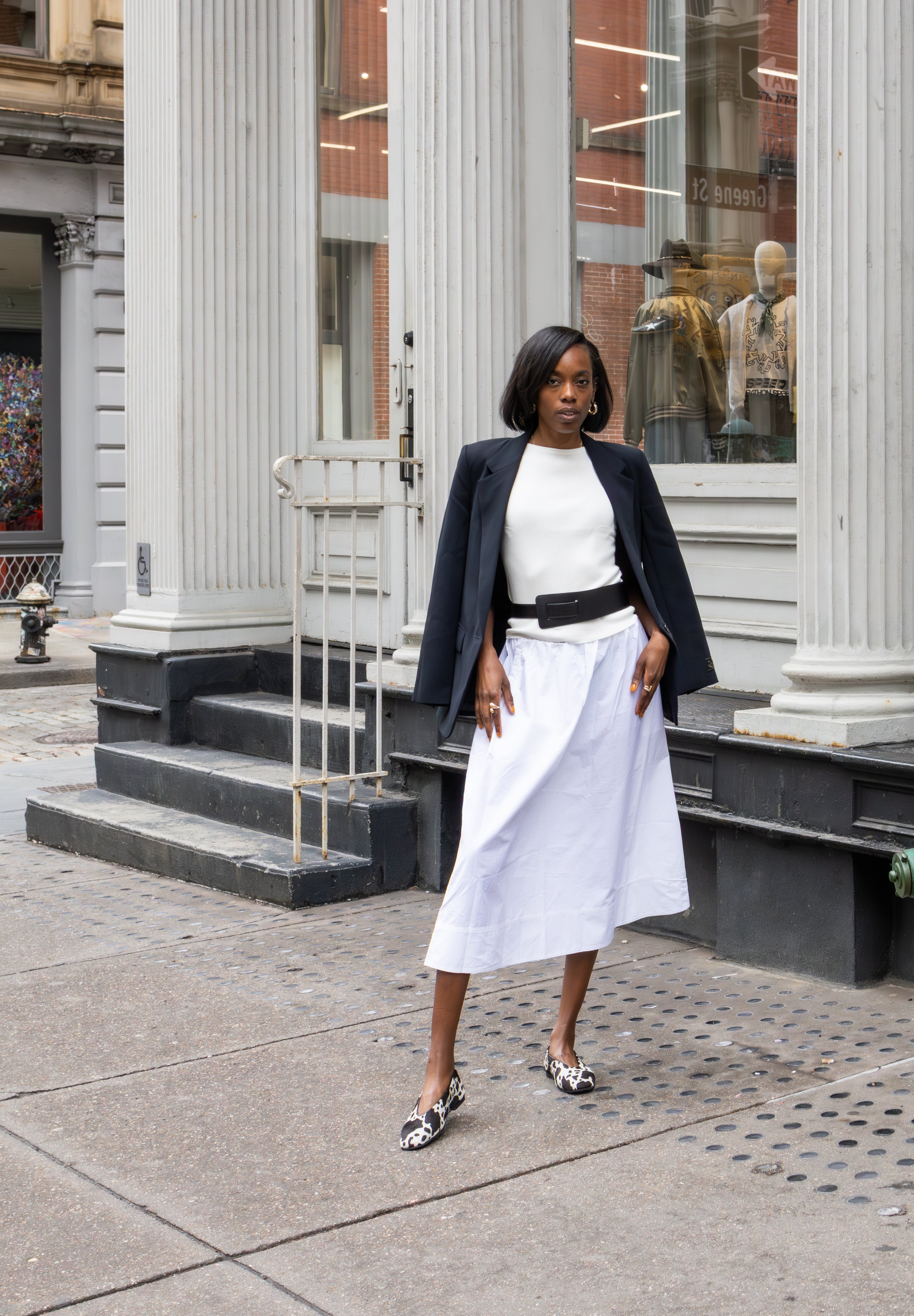 Woman in a white dress and black jacket standing on a city street with classical architecture.